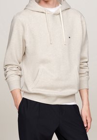Beżowa bluza z kapturem wykonana z miękkiego materiału, z kieszenią kangurką, regulowanym kapturem oraz małym logo na klatce piersiowej. Uzupełniona ciemnymi spodniami.