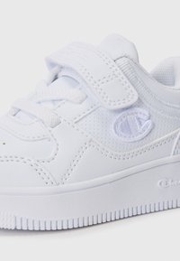 Chaussure de sport blanche avec une tige synthétique texturée, un système de fermeture à lacets et Velcro, et une semelle en caoutchouc avec un motif de semelle.