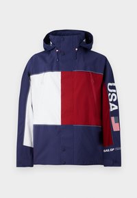 Mørkeblå, rød og hvid sejljakke med hætte, USA tekst og flag på højre ærme, to frontlommer med lynlås og knaplukning.