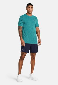 Mann steht da und trägt ein türkisfarbenes, gemustertes Sportshirt, marineblaue Shorts mit Logo, weiße Sneaker sowie sichtbare Tattoos an Armen und Beinen.