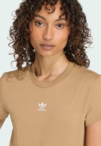 Béžové bavlněné tričko s krátkým rukávem, s bílou vyšitou logem Adidas na hrudi. Klasický kulatý výstřih, měkká textura.