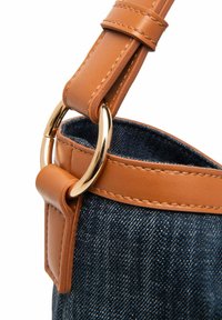 Denimtasche mit einem hellbraunen Lederträger, ausgestattet mit einem goldenen Metallring und einer Schnalle. Sichtbare Nähte entlang des Trägers und eine strukturierte Oberfläche.