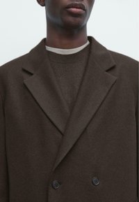 Brązowy płaszcz dwurzędowy z mieszanki wełny z klapami szczytowymi, wyposażony w dwa czarne guziki i miękkie, teksturowane wykończenie. Minimalistyczny design.