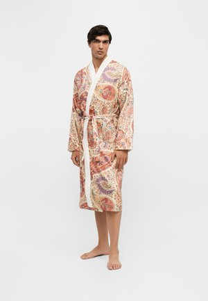 Mann steht barfuß und trägt einen knie-langen Bademantel mit floralem Paisley-Muster, weißem Kragen und Taillengürtel vor einem schlichten Hintergrund.