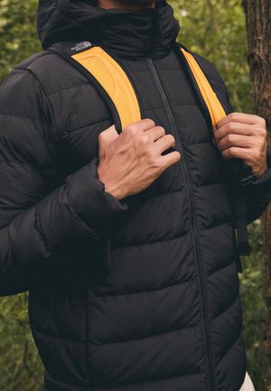 Schwarze Steppjacke mit gesteppter Textur, Reißverschluss und einer angebrachten Kapuze, ergänzt durch gelbe Rucksackriemen.