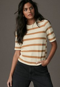 Beige gestreifter Kurzarmpullover mit braunen horizontalen Streifen. Strukturierter Strickstoff mit Rundhalsausschnitt. Getragen mit dunklen Jeans.