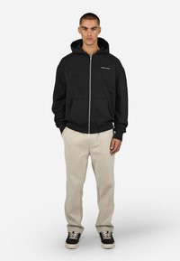 Juodas zip-up gobtuvas, pagamintas iš minkštos medžiagos, turintis priekinę kišenę ir raukšlėtus rankogalius, derinamas su béžinėmis kelnėmis ir juodais sportiniais bateliais.