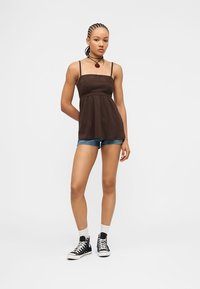 Νέα γυναίκα με πλεξούδες, που φοράει καφέ αμάνικη μπλούζα, τζιν σορτς, μαύρα ψηλά αθλητικά παπούτσια και λευκές κάλτσες, στέκεται με το ένα χέρι πίσω από την πλάτη.