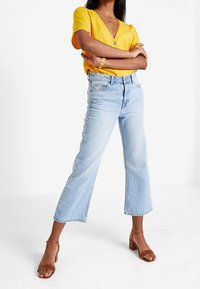 Světle modré, vysoké jeansy s krátkým, rozšířeným designem. Kombinované s živým žlutým topem s výstřihem do V a hnědými sandály na blokovém podpatku.