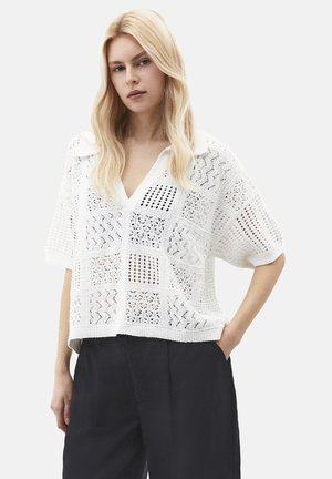 Donna bionda che indossa un maglione bianco a maniche corte in maglia aperta con colletto e pantaloni neri a vita alta, in piedi con una mano in tasca.
