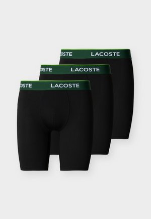 Tre sorte Lacoste boxer briefs med mørkegrønne elastiske linninger og hvide Lacoste-logoer stablet diagonalt på en hvid baggrund.