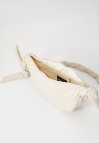 Beige stoffveske med knytthåndtak, glidelås-lukking og glatt tekstur. Innvendig inkluderer en liten glidelåslomme. Minimalistisk design.