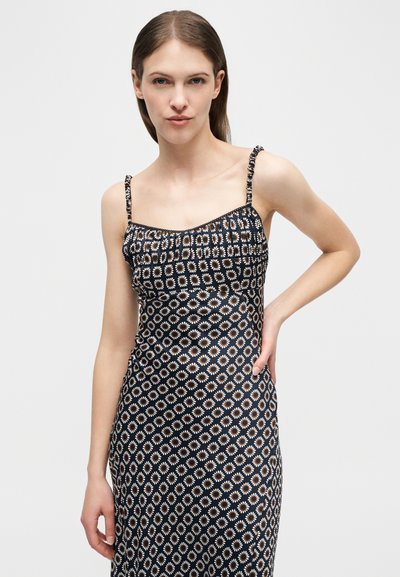 Γυναίκα με ίσια καστανά μαλλιά που φοράει μαύρο αμάνικο φόρεμα με μικρά λευκά και πορτοκαλί λουλούδια, ποζάροντας με το ένα χέρι στη μέση της.