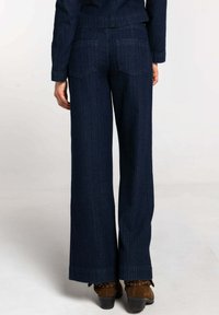 Vaqueros de mezclilla azul oscuro de pierna ancha con bolsillos traseros y detalles de costuras verticales, combinados con botas marrones con adornos de tachuelas.