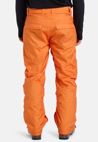 Oranžové snežné nohavice s uvoľneným strihom, ktoré majú dve zadné vrecká, zosilnené švy a hladkú, poveternostne odolnú textúru.
