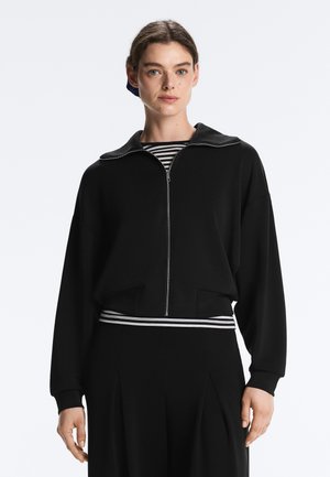 Tânără cu păr închis la culoare poartă o jachetă neagră cu fermoar peste un tricou în dungi alb-negru, stând în fața unui fundal simplu.