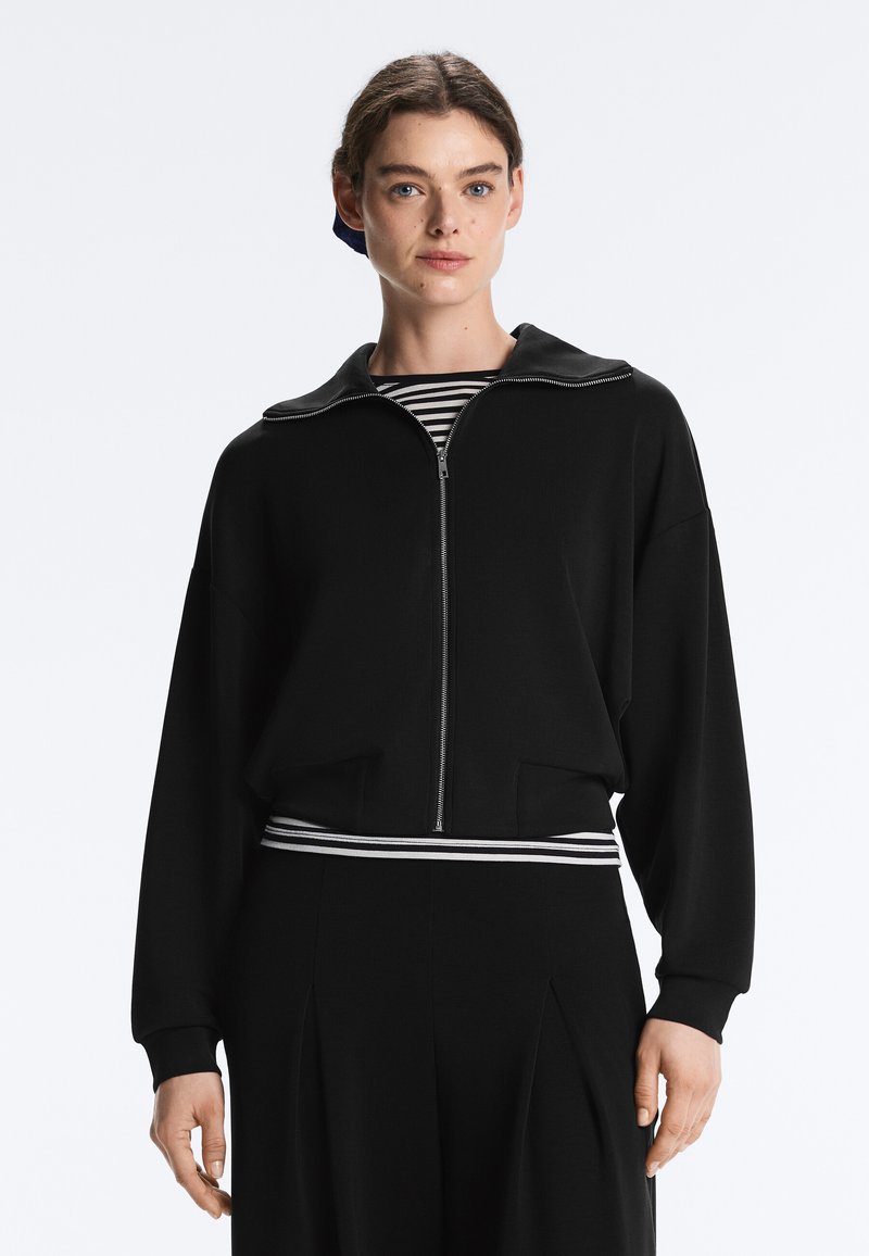 Młoda kobieta o ciemnych włosach nosi czarną kurtkę zapinaną na zamek nałożoną na czarno-białą bluzkę w paski, stoi na jednolitym tle.