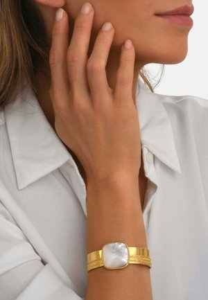 Femme portant un chemisier blanc touche son visage, montrant un bracelet en or avec un grand centre en nacre blanche à son poignet.