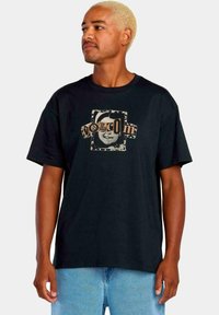 Camiseta de algodón negra con cuello redondo, que presenta un diseño gráfico en beige y gris, que representa una cara y el nombre de la marca "VOLCOM" en el centro.