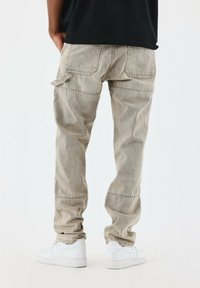 Pantalones cargo de color gris claro con múltiples bolsillos, corte relajado y detalles de costura visibles, combinados con zapatillas blancas. Textura de mezclilla suave.