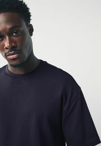 Ung svart man med kort lockigt hår som bär en stor mörk marinblå t-shirt, ser något nedåt och åt sidan, neutral bakgrund.