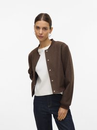 Veste bomber marron courte avec boutons-pression et poignets côtelés, portée sur une chemise blanche, associée à un jean bleu marine.