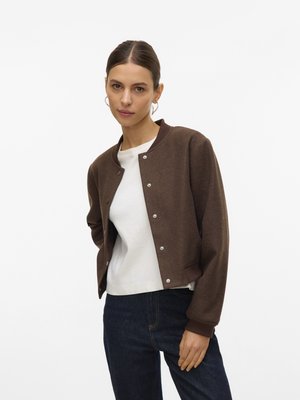 Veste bomber marron courte avec boutons-pression et poignets côtelés, portée sur une chemise blanche, associée à un jean bleu marine.