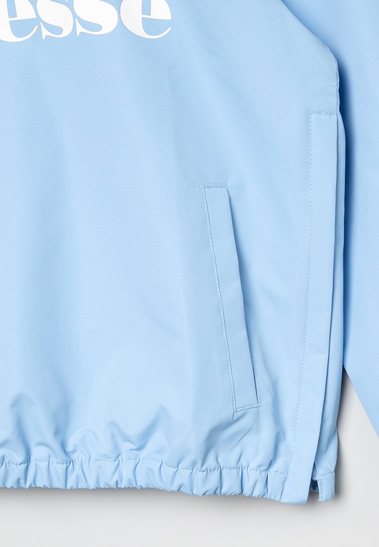 Chaqueta azul clara con dobladillo elástico y bolsillo lateral vertical, logo de la marca parcialmente visible en blanco en la parte superior izquierda.