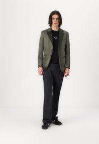 Olivenfarbener Blazer mit satinierter Oberfläche, schwarzes Hemd darunter, dunkle Hose und schwarze Sneaker. Slim Fit mit kontrastierenden Revers.
