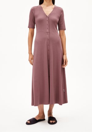 Kobieta w mauve midi sukience zapinanej na guziki z krótkimi rękawami i czarnych, otwartych kapciach stoi na białym tle.