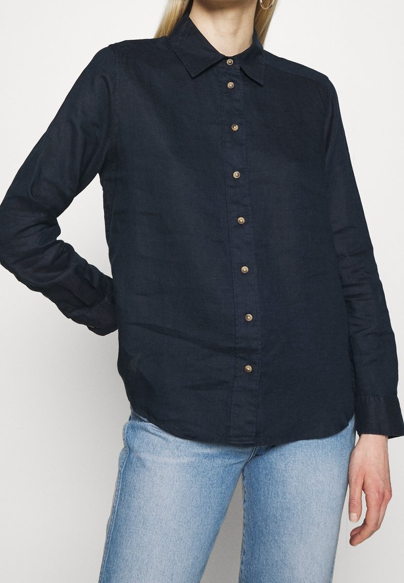 Femme portant une chemise noire à manches longues boutonnée avec un jean bleu clair, debout avec une main sur la hanche devant un fond uni.