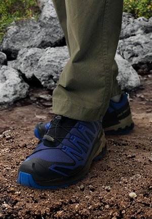 Zapatillas de trail con un diseño en negro y azul, materiales de malla y goma, superficie texturizada y sistema de cordones elásticos, utilizadas en terreno rocoso.