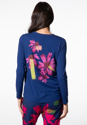 Woman with long dark hair wearing a blue long-sleeve top and pants, both featuring large pink and green floral prints on the back.
