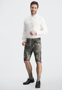 Mann trägt ein weißes Leinenhemd, graue traditionelle Shorts mit blauen Akzenten und knielange graue Socken, kombiniert mit schwarzen Schuhen.