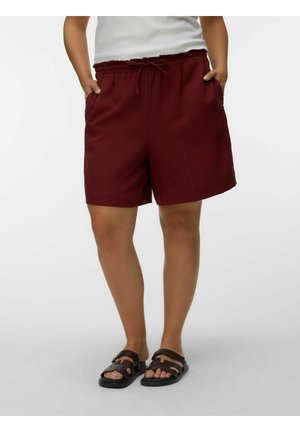 Person iført mørkerøde shorts med snøre og sorte sandaler med åben tå, stående med hænderne i lommerne mod en hvid baggrund.