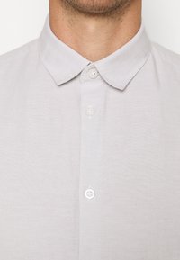 Camicia di lino grigio chiaro con collo button-down e bottoni bianchi lungo la patta frontale. Il tessuto ha un aspetto morbido e strutturato.