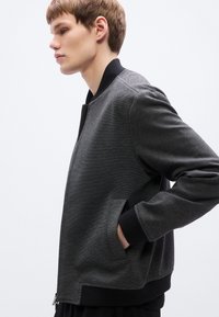 Γκρι τζάκετ μπόμβερ με υφή, μαύρα ριμπ μανσέτα και γιακά, και πλαϊνές τσέπες, με δομημένη εφαρμογή.