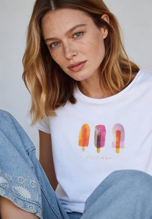 Young woman with light brown hair wearing a white T-shirt featuring three colorful popsicle illustrations and blue jeans with embroidered details.