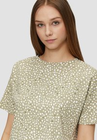 Koszula z krótkim rękawem w jasnym oliwkowym kolorze, z teksturowanym wzorem białych nieregularnych kropek, okrągłym dekoltem i luźnym krojem.