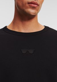 Fekete pamut póló minimalista, dombornyomott napszemüveg grafikával, alatta a "KARL LAGERFELD" felirattal. Klasszikus környakú kivitel.