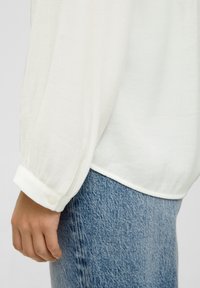 Chemise blanche à manches longues texturée, avec des manches à poignets et des détails de boutons, portée avec un jean en denim bleu. Accent sur le tissu et les éléments de design.