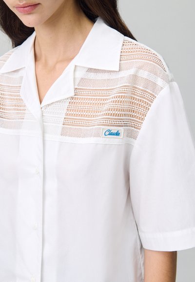 Camicia bianca a maniche corte con un pannello in pizzo testurizzato sul petto e una piccola etichetta blu "Claude" sul lato sinistro. Tessuto liscio.