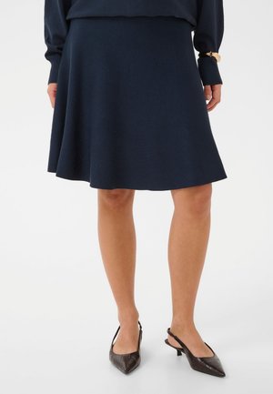 Un mannequin porte une jupe évasée bleu marine arrivant aux genoux, avec un pull assorti et des escarpins foncés à bout pointu et bride arrière.