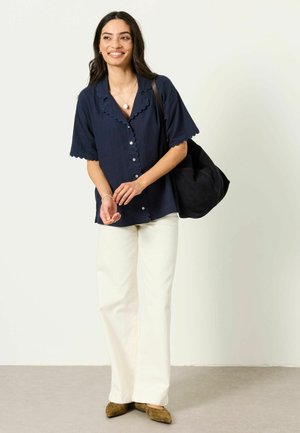 Woman with long dark hair wearing a navy scalloped short-sleeve blouse, white pants, brown shoes, and carrying a black shoulder bag.