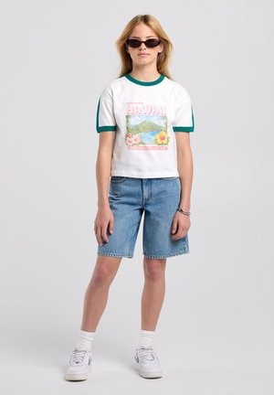 Jonge persoon met zonnebril, wit Hawaii grafisch T-shirt met groene bies, denim shorts, witte sokken en witte sneakers, staand tegen een effen achtergrond.