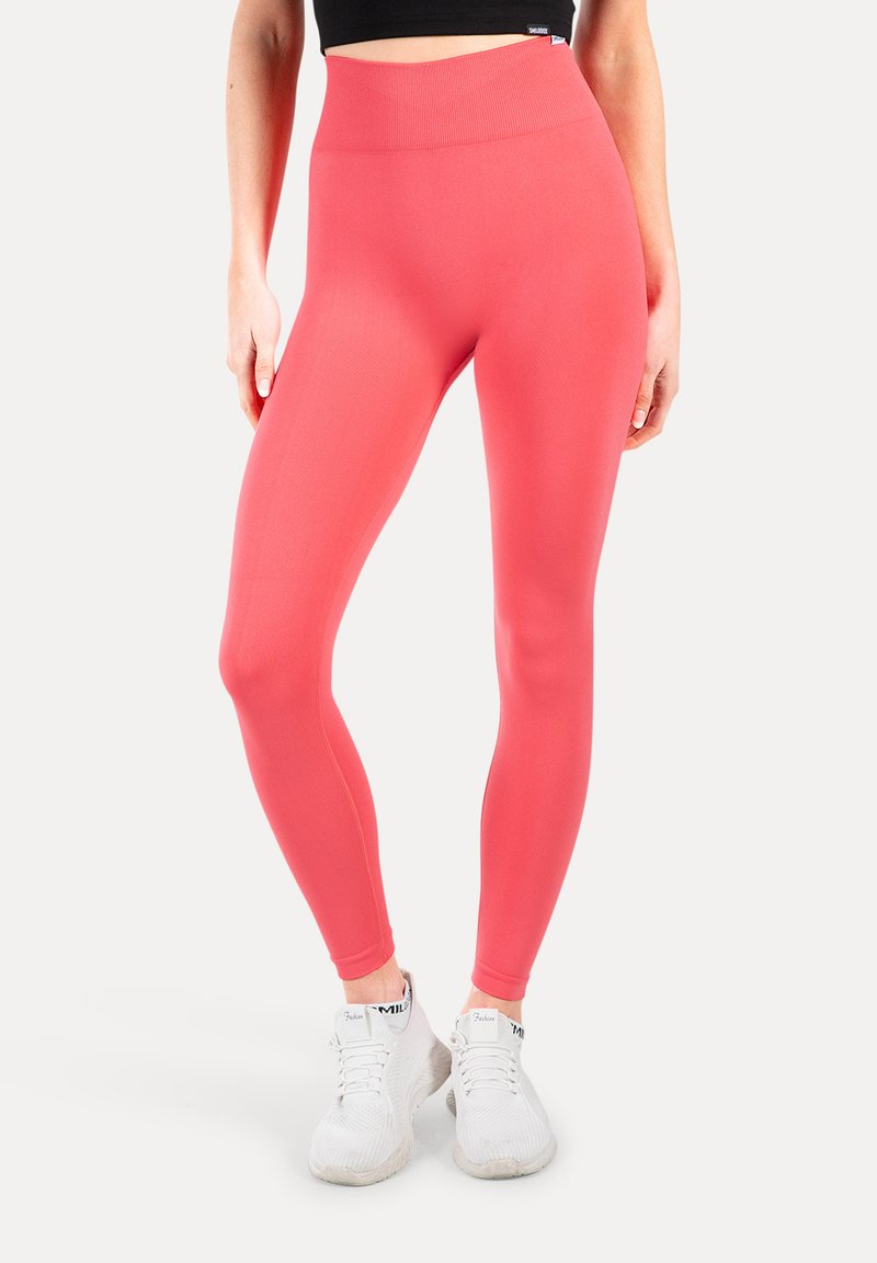 Vrouw die koraalroze leggings met hoge taille en witte sportschoenen draagt, staand met ontspannen armen naast het lichaam tegen een effen achtergrond.