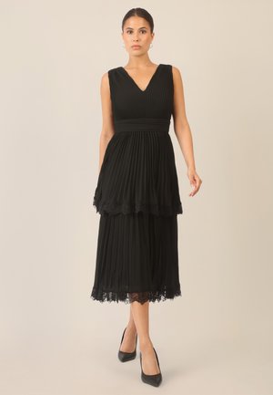 Schwarzes plissiertes Kleid mit V-Ausschnitt, geschichteter Gestaltung und Spitzenbesatz am Saum. Dazu werden schwarze High Heels getragen. Glatte Stofftextur.