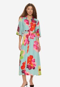 Femme portant une robe midi bleu clair boutonnée avec de grandes imprimés floraux multicolores, taille cintrée et manches jusqu'aux coudes.