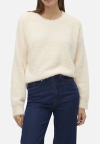 Pull léger en crème, duveteux, avec un col rond et des manches longues, associé à un jean en denim bleu foncé. Texture douce avec une légère complexité.