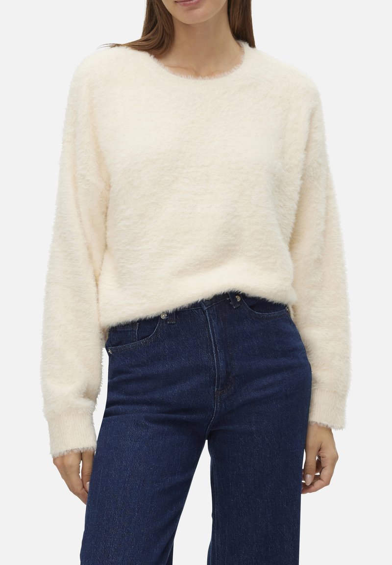 Pull léger en crème, duveteux, avec un col rond et des manches longues, associé à un jean en denim bleu foncé. Texture douce avec une légère complexité.